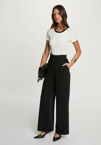 Top bianco aderente con scollatura nera, pantaloni neri a vita alta e gamba larga, borsa nera e décolleté nere a punta con accenti dorati.