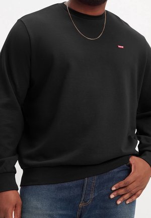 Man in een zwart Levi's sweatshirt, blauwe jeans, gouden ketting om zijn hals, met zijn hand op zijn heup leunend tegen een witte achtergrond.
