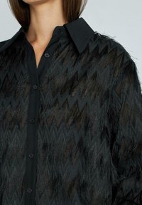 Camisa negra semitranslúcida abotonada con patrones de zigzag y detalles de flecos texturizados, usada por una persona, mostrando solo el cuello y la parte superior del torso.