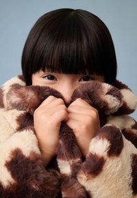 Kurtka z fałszywego futra w beżowo-brązowy wzór w cętki, z wysokim kołnierzem. Ręce chwytają miękki materiał blisko twarzy.