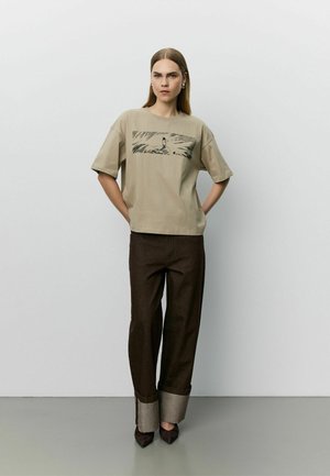 Femme aux cheveux raides portant un t-shirt graphique beige, un pantalon large foncé avec de grands revers, et des chaussures sombres pointues, debout devant un mur uni.