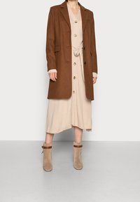 Cappotto di lana marrone con revers, bottoni frontali e tasche laterali, indossato sopra un vestito beige a costine e abbinato a stivaletti beige alla caviglia.