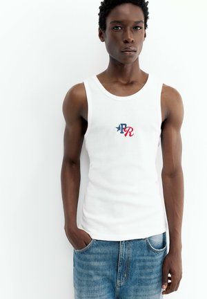 Ung mand med mørk hud iført en hvid ærmeløs trøje med blåt og rødt logo og blå jeans, står med den ene hånd i lommen.