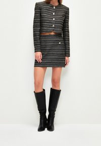 Vrouw in een zwart-wit gestreepte cropped jack met knopen, bijpassende minirok en zwarte kniehoge laarzen.