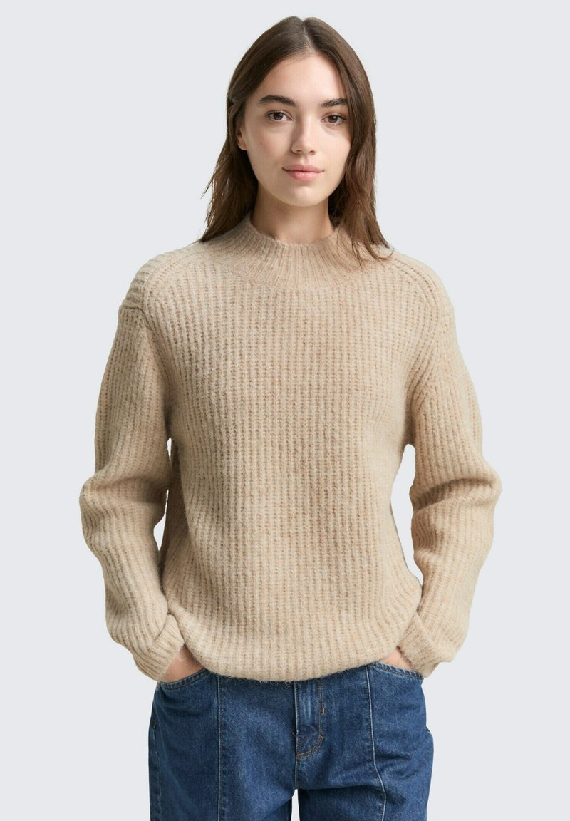 Beige gestrickter Pullover mit langen Ärmeln und hohem Halskragen, der ein strukturiertes Rippmuster aufweist. Kombiniert mit blauen Jeans.