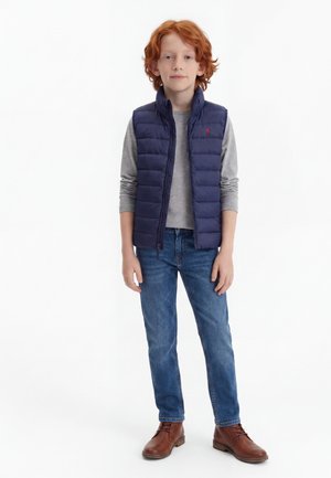 Κοκκινομάλλης αγόρι όρθιο, φοράει μπλε σκούφο γιλέκο πάνω από γκρι μακρυμάνικο μπλουζάκι, μπλε τζιν και καφέ μπότες με κορδόνια σε λευκό φόντο.