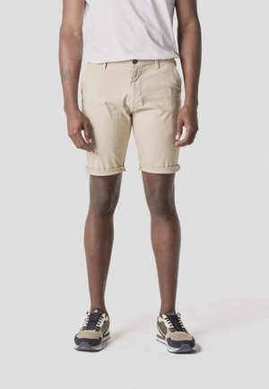 Hombre vestido con pantalones cortos beige remangados, camiseta blanca y zapatillas verde oliva, de pie contra un fondo liso.