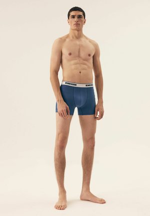 Herren Boxerbriefs in Marineblau mit einem weißen elastischen Bund, auf dem "HENDERSON" aufgedruckt ist. Der Stoff wirkt glatt und eng anliegend. Model steht barfuß.