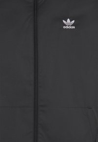 Černá bunda vpředu se středním zipem a bílým vyšívaným logem Adidas trefoil na levé hrudi.