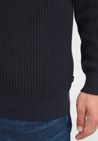 Dunkelblauer Strickpullover mit geripptem Saum und Bündchen, der strukturierte Nähte und ein kleines Etikett an der Seite aufweist. Sichtbare Jeans aus Denim.