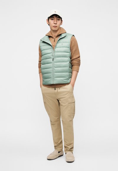 Μπουφάν σε απόχρωση μέντας με φερμουάρ, φορεμένο πάνω από καφέ πουλόβερ με υφή. Συνδυασμένο με μπεζ παντελόνι cargo και μπεζ loafers, ολοκληρωμένο με λευκό καπέλο.