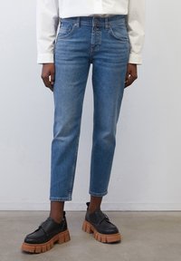 Jeans de denim azul claro con un ajuste entallado, diseño de cinco bolsillos y detalles cosidos, combinados con zapatos negros y una camisa blanca.