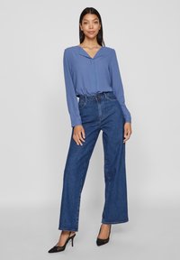 Donna in piedi con una blusa azzurra a maniche lunghe, jeans in denim a vita alta e gamba larga, e tacchi a punta neri su uno sfondo neutro.