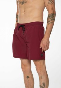 Shorts de bain bordeaux avec une taille élastique et un cordon de serrage, poches latérales, et un petit logo circulaire sur l'ourlet.
