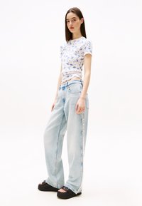 Tricou cu imprimeu floral, cu mâneci scurte, asortat cu blugi largi de culoare bleu deschis. Modelul poartă sandale cu platformă negre cu catarame. Fundal neutru.