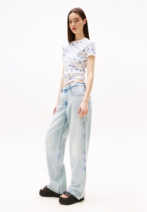 T-shirt à imprimé floral avec des manches courtes, associé à un jean évasé bleu clair. Le modèle porte des sandales compensées noires avec des boucles. Fond neutre.