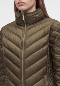 Femme portant une veste matelassée vert olive avec un col montant et des détails visibles de fermeture éclair dorés, montrée des épaules au menton.