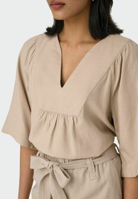 Beige Leinenoberteil mit tiefem V-Ausschnitt, Puffärmeln und gerafftem Detail an der Taille. Akzentuiert mit einem passenden Stoffgürtel.