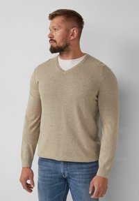 Beiger V-Ausschnitt-Pullover aus weichem Stoff mit langen Ärmeln, kombiniert mit blauen Jeans. Leichte Textur, figurnahes Design.