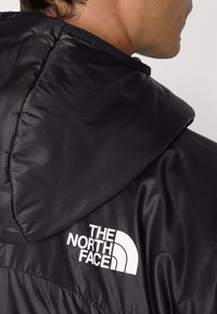 Zwarte hoodie gemaakt van gladde stof, met een wit logo op de achterkant. Het ontwerp bevat naaddetails en een gestructureerd oppervlak.