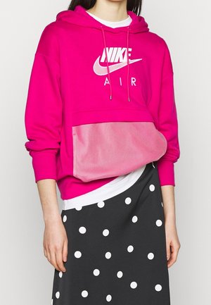 Osoba nosząca jaskraworóżową bluzę Nike Air z kieszenią kangura i czarną spódnicę w białe grochy, stojąca na jednolitym jasnym tle.