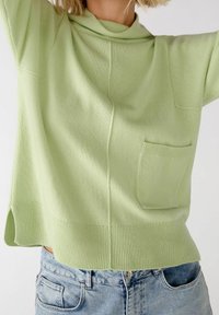 Donna che indossa un maglione lavorato a maglia verde chiaro con tasca frontale e jeans di denim blu chiaro, braccia sollevate sopra la testa, sfondo semplice.