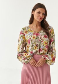 Blusa floral de manga larga en color crema, con flores rosas y amarillas, combinada con una falda plisada en tono rosa claro. Detalle de tejido texturizado visible.