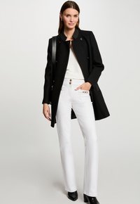 Cappotto nero su misura con colletto alto, indossato sopra un top bianco. Abbinato a jeans bianchi a gamba dritta con dettagli dorati e tacchi neri.