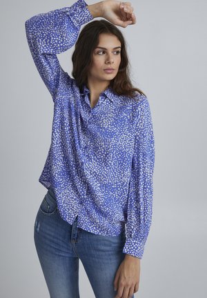 Blaues Hemd mit Button-Down-Kragen, weißen Polka-Dots, langen Ärmeln und lockerer Passform. Kombiniert mit blauen Jeans.