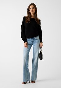 Zwarte gebreide trui met off-shoulder ontwerp, gecombineerd met lichtblauwe high-waisted wijde broekspijpen jeans. Accessoires omvatten een kleine zwarte tas.
