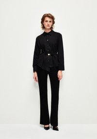 Camicia nera con cintura, colletto e chiusura con bottoni, abbinata a pantaloni neri a campana e décolleté con punta. Tessuto liscio.