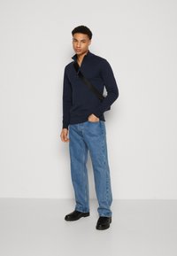 Camiseta de manga larga azul marino con cremallera, jeans rectos de mezclilla azul, zapatos de suela de goma negros, modelo de pie con una postura casual y bolsa de hombro.