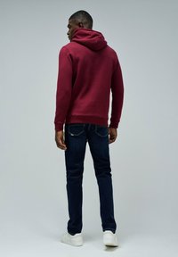 Mężczyzna odwrócony plecami, ubrany w bordową bluzę z kapturem, ciemnoniebieskie dżinsy i białe trampki na jednolitym szarym tle.