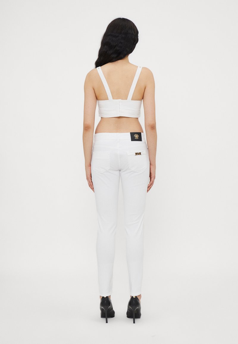 Witte, strakke jeans en een bijpassend cropped top, beide gemaakt van een gladde stof, met dunne bandjes en een logo-patch op de rug. Zwarte hakken.