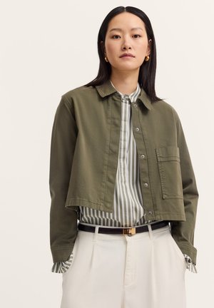 Olivgrüne, kürzere Jacke mit Knopfleiste und einer Brusttasche. Getragen über einem grauen gestreiften Hemd und kombiniert mit cremefarbener Hose.