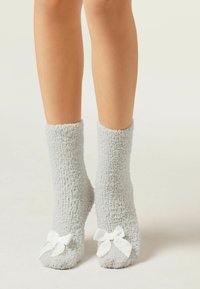 Graue, flauschige Socken mit weicher Textur, versehen mit weißen Schleifen aus Satin an der Spitze, in Knöchelhöhe. Gemütliches und warmes Design für den Innenbereich.