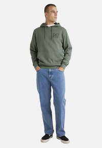 Green hoodie with a front pocket and drawstrings, paired with blue denim jeans. Black slip-on shoes, minimal patterns, and a relaxed fit.