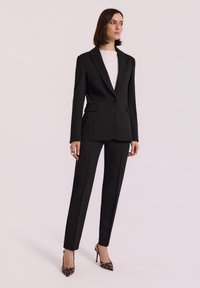 Kobieta w czarnym, dopasowanym żakiecie i spodniach, białej bluzce, wysokich szpilkach w leopardzi wzór i dużym pierścionku, stojąca na jednolitym jasnym tle.