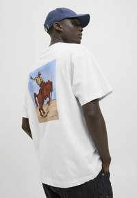 Camiseta de algodón blanca con una ilustración colorida de un vaquero y un caballo en la parte posterior, con escote redondo y mangas cortas.