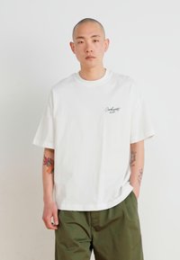 Camiseta de algodón blanca de manga corta, corte holgado, cuello redondo y un pequeño logotipo bordado en el lado izquierdo del pecho.