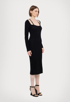 Black long-sleeve midi dress with ribbed texture, featuring adjustable straps. Paired with black heeled sandals and gold accents.