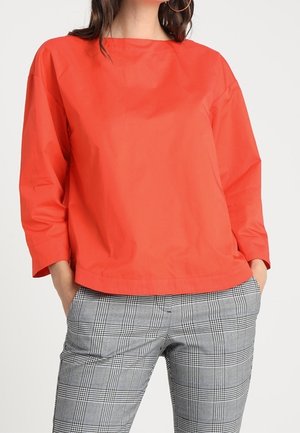 Vrouw draagt een feloranje top met lange mouwen en grijze geruite broek, met handen in de zakken, zichtbaar van nek tot heupen.