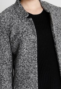 Chaqueta de mezclilla de lana texturizada y gris con cuello, de frente abierto y mangas largas, superpuesta sobre un suéter de punto negro.