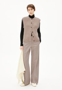 Beige gebreid vest met een ribbelstructuur, knoopsluiting aan de voorkant, gedragen over een zwarte turtleneck. Grijze op maat gemaakte broek en een crèmekleurige sjaal maken de outfit compleet.