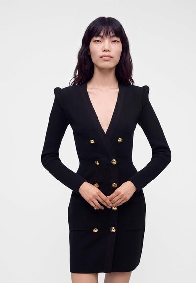 Femme aux longs cheveux foncés portant une robe noire ajustée avec des boutons dorés, debout devant un fond clair uni, les mains jointes.