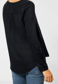Czarna bluzka z długim rękawem, z plisowanym tyłem, gładką fakturą, zapinanymi mankietami i zaokrąglonym dołem, w połączeniu z niebieskimi dżinsami.