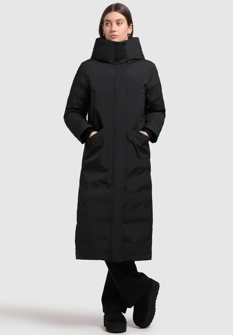 Sort lang pufferjakke med høj krave, frontzipper og sidelommer. Lavet af glat, polstret materiale. Båret med sorte bukser og støvler.