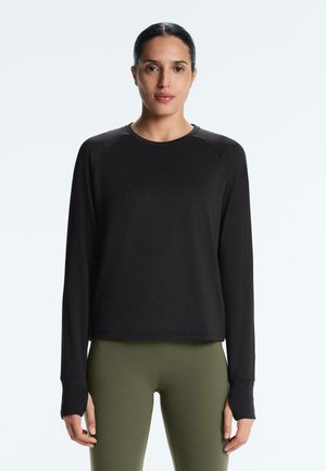 Kvinde med mørkt hår iført en sort langærmet top med tommelfingerhuller og olivengrønne leggings, vendt fremad mod en ensartet hvid baggrund.