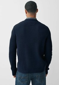Mørkeblå strikket sweater med et struktureret ribdesign, der har høj krave og tætsiddende manchetter. Kombination med lyseblå jeans.
