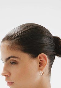 Pendientes de aro en oro rosa con un diseño de triple banda, que presentan un acabado suave y una ligera curvatura, llevados en un peinado elegante.